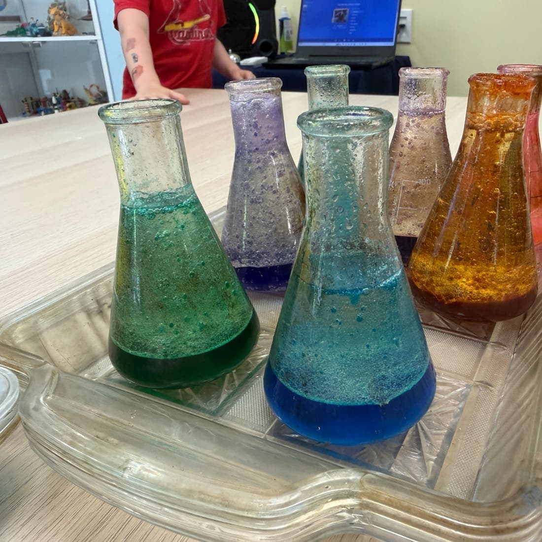 Six flasks of bubbling coloured liquid lined up on a tray during a camp chemistry experiment.